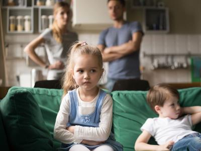 Two children look sad sat on sofa while parents stand in background